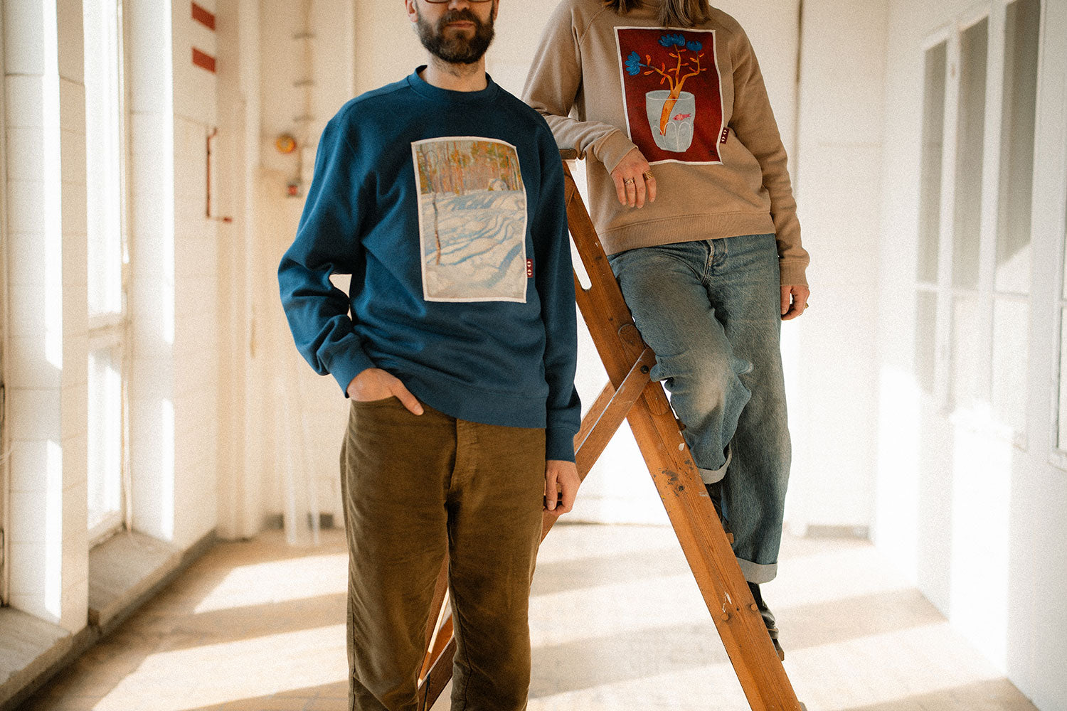 Two people standing on a wooden ladder wearing graphic sweatshirts in a bright room.