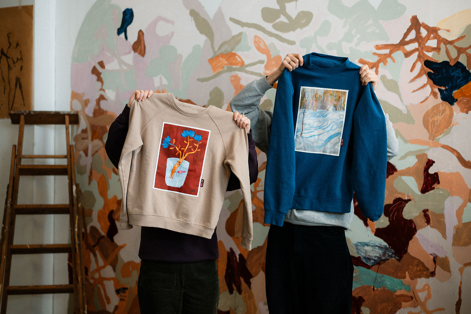 Two people holding up a beige sweatshirt and a blue sweatshirt against a colorful wall.
